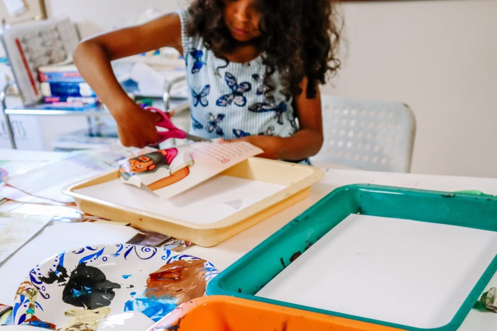 Little girl sits at white table to cut out pictures of a magazine to make a craft