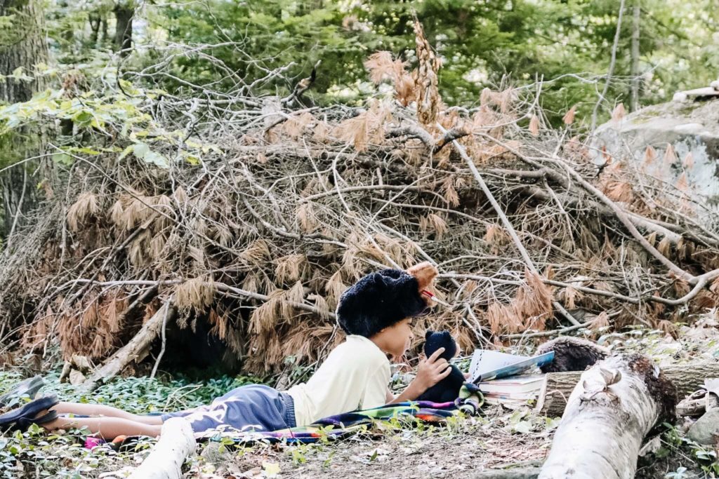 Little boy lays down outside with teddy bear to read a book int he woods