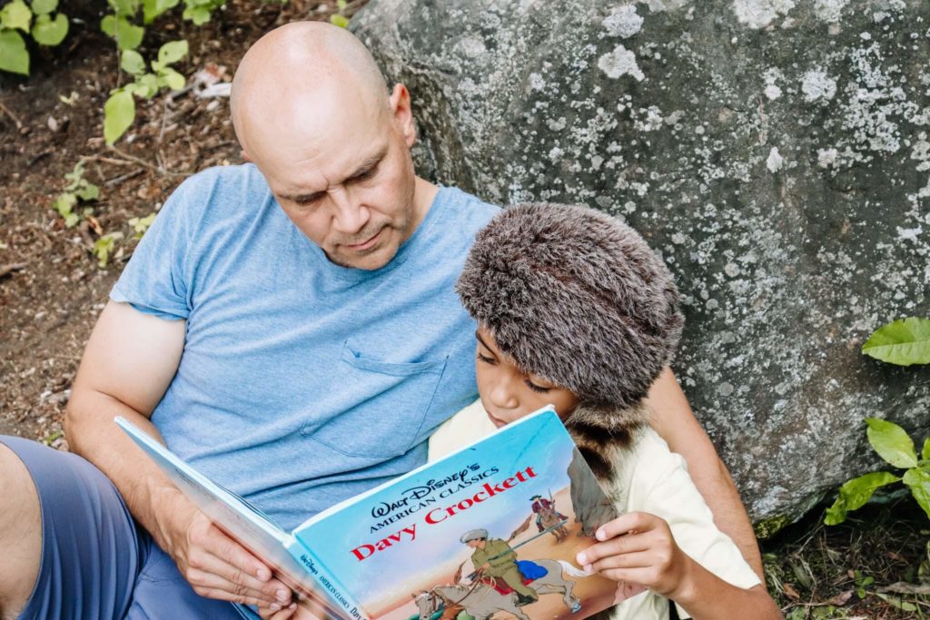 Man reading book aloud  to young son against a rock